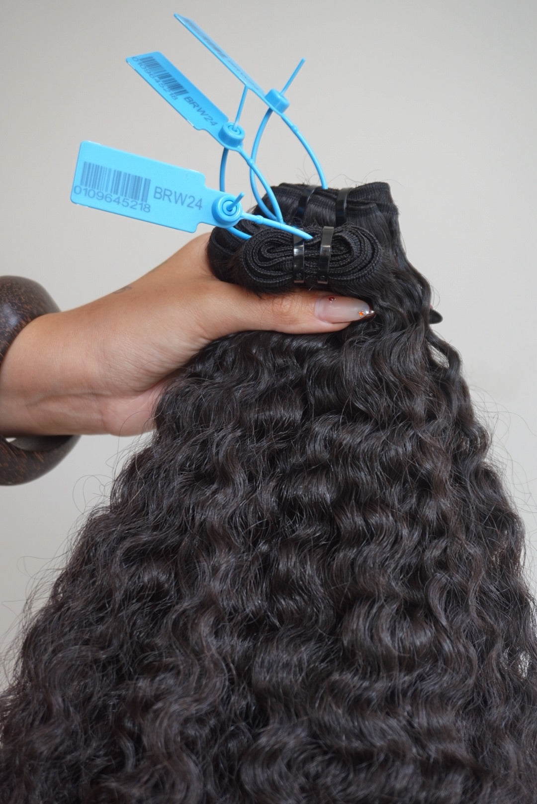 Curly hair extensions held by a hand with blue clips on a neutral background