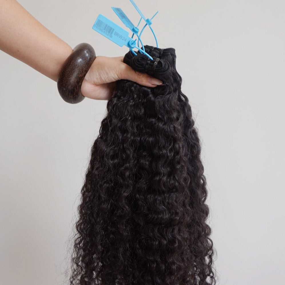 Black curly hair extensions held by a hand with a wooden bangle on a light gray background